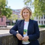 Die rheinland-pfälzische Umweltministerin Katrin Eder steht im Freien vor ihrem Ministerium in Mainz. Sie trägt einen dunkelblauen Blazer und ein weißes T-Shirt mit Blumenmotiv. Im Hintergrund sind Bäume, ein Platz und Verwaltungsgebäude zu sehen.