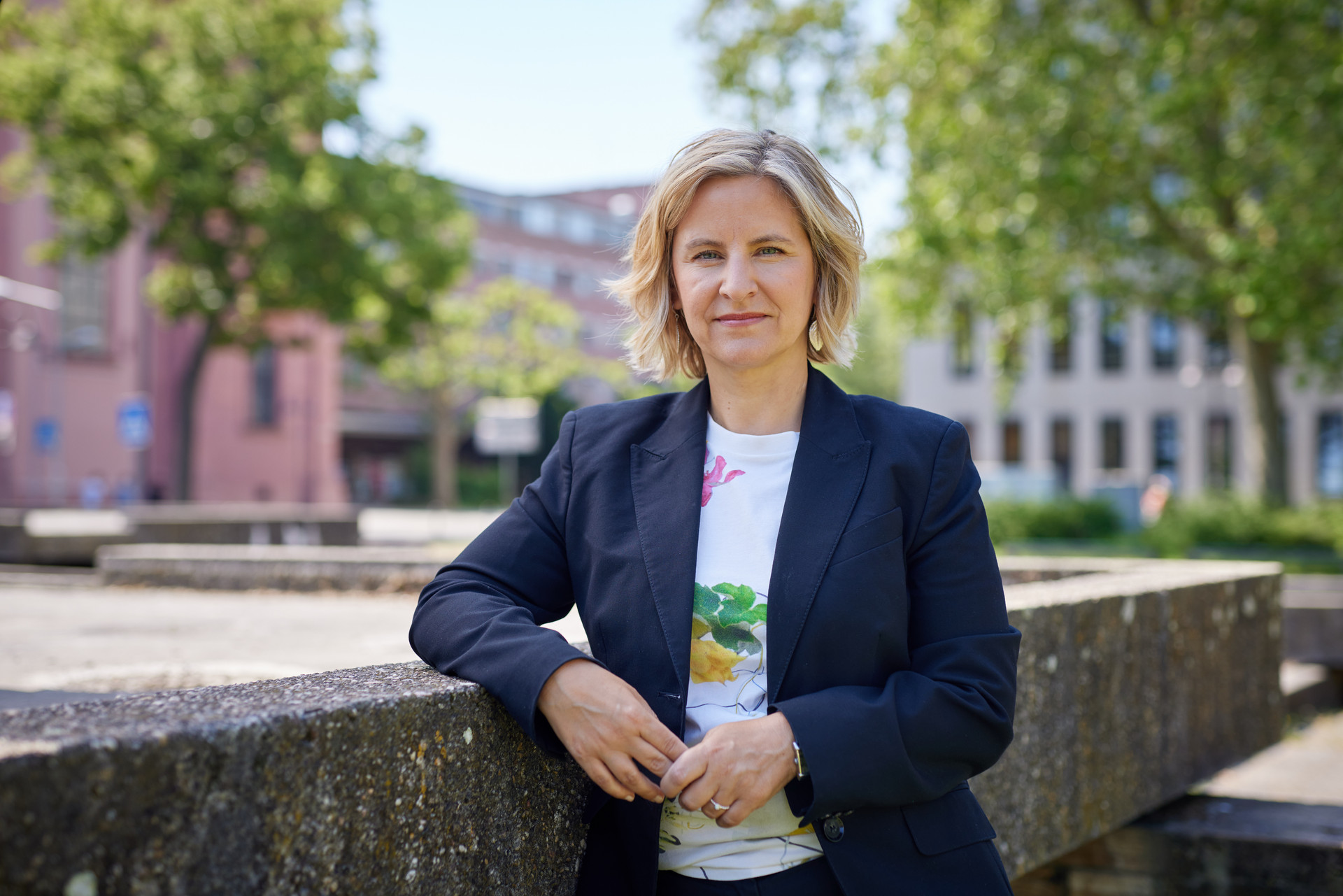 Die rheinland-pfälzische Umweltministerin Katrin Eder steht im Freien vor ihrem Ministerium in Mainz. Sie trägt einen dunkelblauen Blazer und ein weißes T-Shirt mit Blumenmotiv. Im Hintergrund sind Bäume, ein Platz und Verwaltungsgebäude zu sehen.