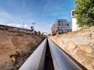 Zwei große Fernwärmerohre werden in einem aufgeschütteten Graben entlang einer Straße verlegt, im Hintergrund sind Bauarbeiten und Gebäude zu sehen.