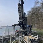 Bohrgerät bei der Verlegung von Erdsonden für ein kaltes Nahwärmenetz: Auf einer Baustelle sind gestapelte Sondenrohre zu sehen, während ein Arbeiter die Tiefenbohrung zur Nutzung von Erdwärme vorbereitet.