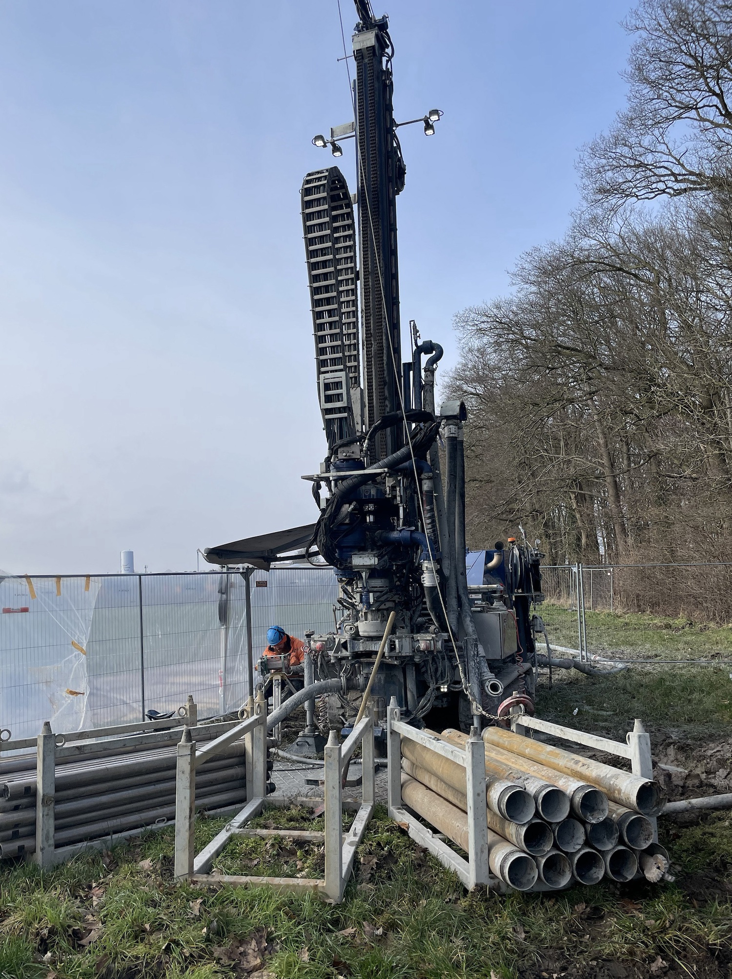 Bohrgerät bei der Verlegung von Erdsonden für ein kaltes Nahwärmenetz: Auf einer Baustelle sind gestapelte Sondenrohre zu sehen, während ein Arbeiter die Tiefenbohrung zur Nutzung von Erdwärme vorbereitet.