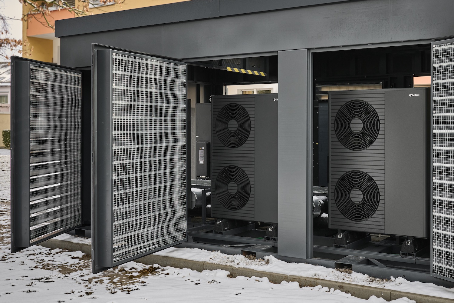 Sechsstufige Wärmepumpenanlage „Energy Unit 2.0“ in der XL-Variante: Außen aufgestellte Luft/Wasser-Wärmepumpenmodule in schallgedämmter Einhausung vor einem Mehrfamilienhaus, teilweise mit geöffneten Schutzgittern.