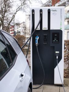 Öffentliche Ladesäule für Elektroautos in einem Wohngebiet, an der ein weißes E-Auto angeschlossen ist und lädt.
