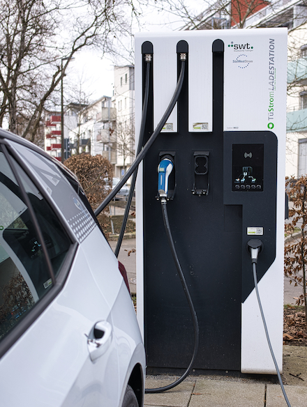 Öffentliche Ladesäule für Elektroautos in einem Wohngebiet, an der ein weißes E-Auto angeschlossen ist und lädt.