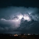 Dunkle Gewitterwolken mit einem hellen Blitz über einer Stadt bei Nacht, im Vordergrund sind Lichter der bebauten Umgebung zu sehen.