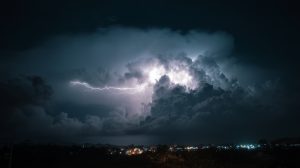 Dunkle Gewitterwolken mit einem hellen Blitz über einer Stadt bei Nacht, im Vordergrund sind Lichter der bebauten Umgebung zu sehen.