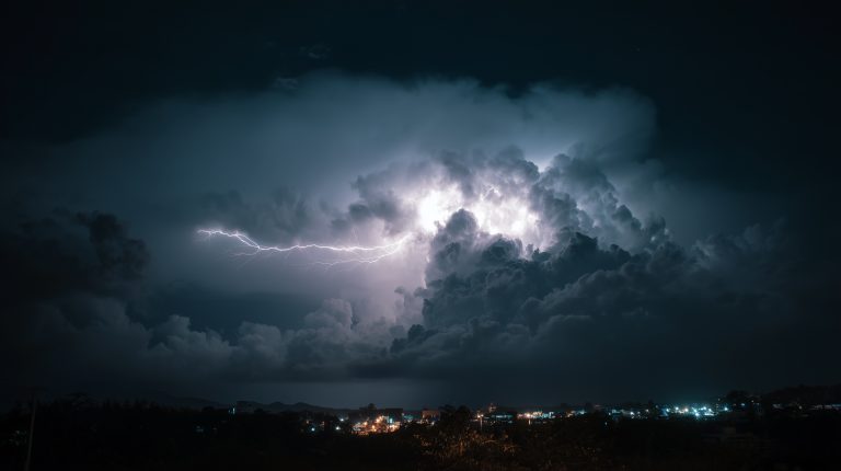 Dunkle Gewitterwolken mit einem hellen Blitz über einer Stadt bei Nacht, im Vordergrund sind Lichter der bebauten Umgebung zu sehen.