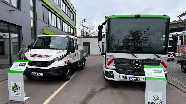 Zwei neue Elektro-Nutzfahrzeuge der Mainzer Entsorgungsbetriebe stehen vor einem modernen Verwaltungsgebäude: links ein weißer Iveco-Transporter mit Ladefläche, rechts ein größerer Mercedes-Benz-Lkw. Vor beiden Fahrzeugen informieren grüne Aufsteller über die Ausstattung und den Einsatz im kommunalen Betrieb.