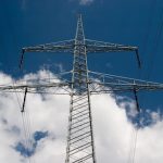 Hochspannungsmast mit mehreren Stromleitungen vor blauem Himmel mit Wolken.