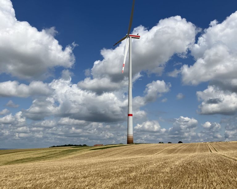 Einzelne Windkraftanlage auf einer leicht hügeligen, abgeernteten Feldfläche. Der hohe Turm mit rot-weißen Markierungen ragt in einen blauen Himmel mit großen, weißen Wolken. Die Landschaft wirkt weit und offen, im Vordergrund erstreckt sich ein goldenes Getreidefeld.