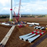 Auf einem weiten Ackerland wird eine Windenergieanlage errichtet: Ein großer Turm steht bereits, ein roter Baukran montiert weitere Bauteile. Am Boden liegen Rotorblätter mit roten Spitzen, Turmsegmente und Technikcontainer, im Hintergrund sind mehrere fertige Windräder in der Landschaft zu sehen.