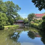 Historische Sägemühle an einem ruhigen Mühlteich, umgeben von Bäumen; Gebäude mit Wasserkraftanlage am Ufer, Spiegelung im Wasser bei sonnigem Wetter.