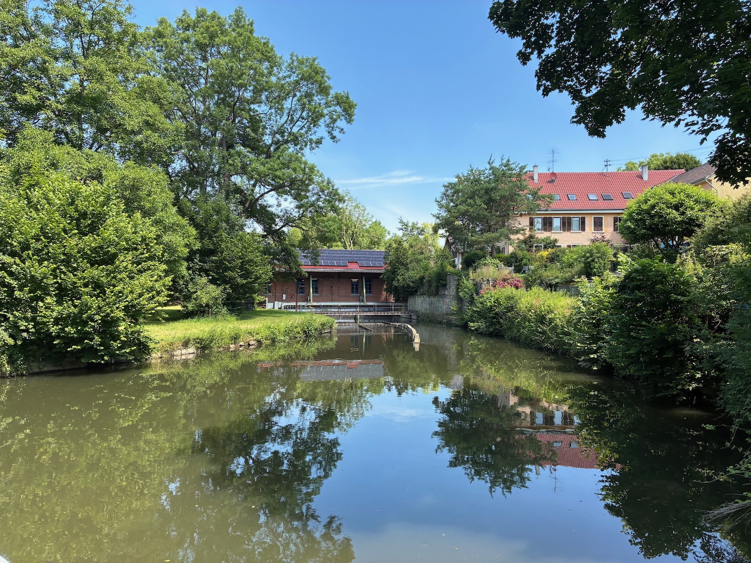Historische Sägemühle an einem ruhigen Mühlteich, umgeben von Bäumen; Gebäude mit Wasserkraftanlage am Ufer, Spiegelung im Wasser bei sonnigem Wetter.