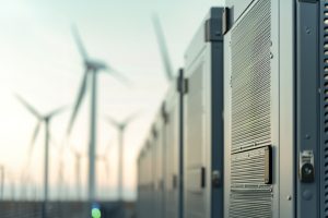Nahaufnahme einer Reihe von Batteriespeicher-Containern in einem Windpark. Die technischen Gehäuse mit Lüftungsgittern stehen in einer Linie im Vordergrund, während im Hintergrund mehrere Windkraftanlagen unscharf zu sehen sind. Die Szene vermittelt die Kombination aus Windenergie und Energiespeicherung.