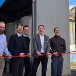 Fünf Männer stehen nebeneinander vor einer technischen Anlage und durchschneiden gemeinsam ein rotes Band. Sie halten Scheren in den Händen. Im Hintergrund sind industrielle Rohrleitungen und ein Container zu sehen, daneben Fahnen mit Unternehmenslogo. Die Szene zeigt eine symbolische Inbetriebnahme im industriellen Umfeld.