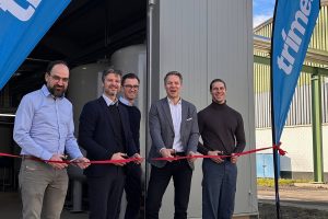 Fünf Männer stehen nebeneinander vor einer technischen Anlage und durchschneiden gemeinsam ein rotes Band. Sie halten Scheren in den Händen. Im Hintergrund sind industrielle Rohrleitungen und ein Container zu sehen, daneben Fahnen mit Unternehmenslogo. Die Szene zeigt eine symbolische Inbetriebnahme im industriellen Umfeld.