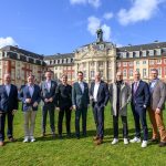 Eine Gruppe von neun Männern steht auf einer Wiese vor einem großen barocken Gebäude mit roter Fassade und heller Verzierung. Die Männer tragen überwiegend Anzüge und schauen in die Kamera, während im Hintergrund das historische Gebäude und ein blauer Himmel mit vereinzelten Wolken zu sehen sind.