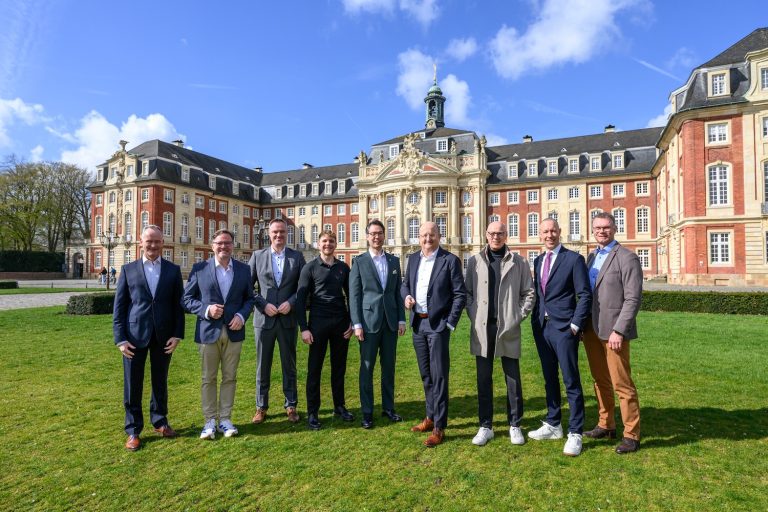Eine Gruppe von neun Männern steht auf einer Wiese vor einem großen barocken Gebäude mit roter Fassade und heller Verzierung. Die Männer tragen überwiegend Anzüge und schauen in die Kamera, während im Hintergrund das historische Gebäude und ein blauer Himmel mit vereinzelten Wolken zu sehen sind.