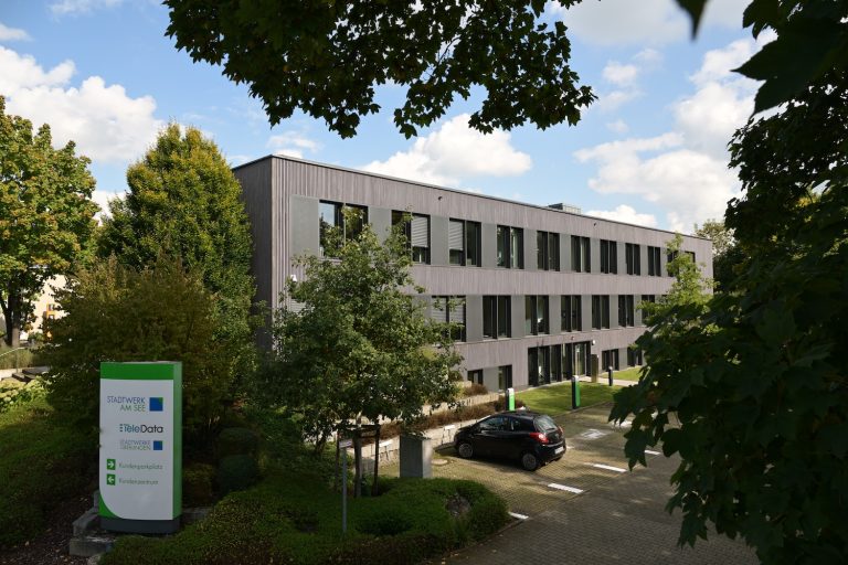 Hauptsitz des Stadtwerks am See in einem modernen, dreigeschossigen Bürogebäude mit grauer Fassade, umgeben von Bäumen und Grünflächen, mit Parkplatz und Firmenschild im Vordergrund.