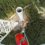 Bau einer Windkraftanlage aus der Vogelperspektive: Ein Kran hebt Turmteile über einem Waldgebiet, Arbeiterinnen und Arbeiter sind am Fundament im Einsatz.