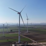 Mehrere moderne Windenergieanlagen stehen im Windpark Minfeld in einer offenen Agrarlandschaft. Die hohen Windräder drehen sich über Feldern und Wegen, im Hintergrund sind weitere Anlagen sowie eine leicht hügelige Landschaft zu erkennen.
