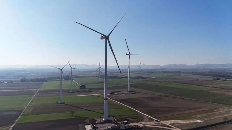 Mehrere moderne Windenergieanlagen stehen im Windpark Minfeld in einer offenen Agrarlandschaft. Die hohen Windräder drehen sich über Feldern und Wegen, im Hintergrund sind weitere Anlagen sowie eine leicht hügelige Landschaft zu erkennen.