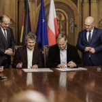 Mehrere Männer stehen und sitzen an einem großen Holztisch in einem repräsentativen Raum und unterzeichnen gemeinsam Dokumente. Zwei Männer sitzen in der Mitte und unterschreiben die Absichtserklärung, während andere Beteiligte daneben stehen und zuschauen. Im Hintergrund sind die Flaggen Deutschlands, der Europäischen Union und Polens zu sehen.