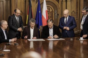 Mehrere Männer stehen und sitzen an einem großen Holztisch in einem repräsentativen Raum und unterzeichnen gemeinsam Dokumente. Zwei Männer sitzen in der Mitte und unterschreiben die Absichtserklärung, während andere Beteiligte daneben stehen und zuschauen. Im Hintergrund sind die Flaggen Deutschlands, der Europäischen Union und Polens zu sehen.