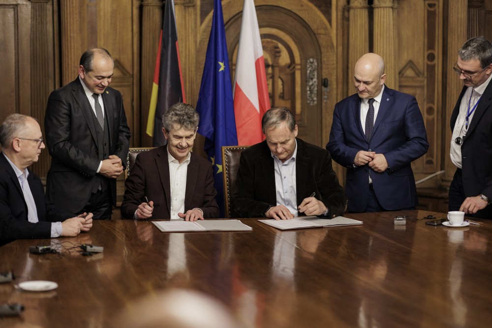 Mehrere Männer stehen und sitzen an einem großen Holztisch in einem repräsentativen Raum und unterzeichnen gemeinsam Dokumente. Zwei Männer sitzen in der Mitte und unterschreiben die Absichtserklärung, während andere Beteiligte daneben stehen und zuschauen. Im Hintergrund sind die Flaggen Deutschlands, der Europäischen Union und Polens zu sehen.