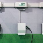 Drei weiße Wallboxen für Elektroautos sind an einer Innenwand montiert und über Kabelkanäle mit Sicherungskästen verbunden. An der mittleren Ladestation steckt ein Ladekabel. Die Wand ist grau und im unteren Bereich grün gestrichen.