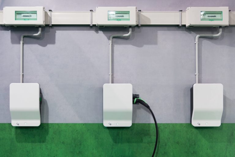 Drei weiße Wallboxen für Elektroautos sind an einer Innenwand montiert und über Kabelkanäle mit Sicherungskästen verbunden. An der mittleren Ladestation steckt ein Ladekabel. Die Wand ist grau und im unteren Bereich grün gestrichen.