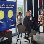 Vier Personen sitzen bei einer Podiumsdiskussion in einem Veranstaltungsraum. Eine Person spricht mit Mikrofon, daneben hört eine Moderatorin zu. Auf großen Bildschirmen sind Informationen zu einem Solarparkprojekt zu sehen, darunter „51,8 GWh jährliche Stromerzeugung“ und „14.000 Haushalte jährlich mit Strom versorgt“. Im Hintergrund fotografiert eine Person die Runde.