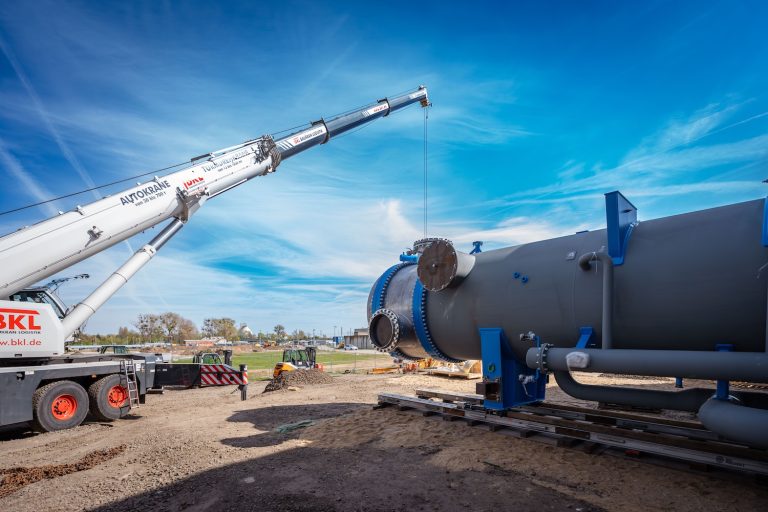 Ein großer Autokran hebt auf einer Baustelle einen zylinderförmigen Industriebehälter aus Metall an. Das Bauteil liegt auf Schienen und ist mit mehreren Rohranschlüssen versehen. Im Hintergrund sind Baugeräte, Fahrzeuge und freie Flächen zu sehen. Darüber spannt sich ein blauer Himmel mit dünnen Wolken.