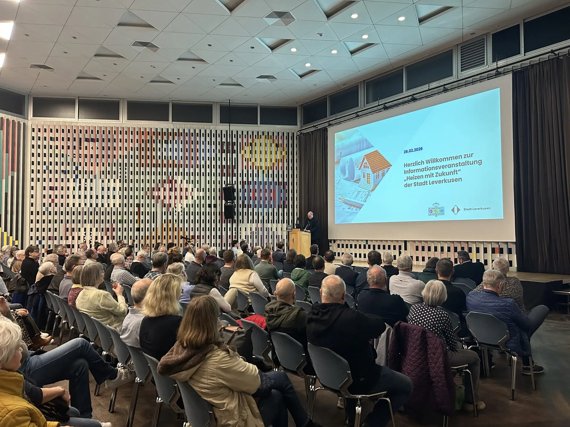 Alt-Text: Bürgerversammlung der Stadt Leverkusen zur kommunalen Wärmeplanung: Zahlreiche Bürgerinnen und Bürger sitzen in einem Veranstaltungsraum und verfolgen einen Vortrag auf einer großen Leinwand, auf der „Heizen mit Zukunft“ präsentiert wird.