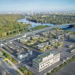 Luftaufnahme einer modernen Busanlage der BVG in Berlin: Auf einem großflächigen Betriebshof stehen zahlreiche gelbe Elektrobusse in Reih und Glied an Ladeplätzen unter überdachten Infrastrukturen. Mehrere Funktionsgebäude mit begrünten Dächern und Solaranlagen umgeben das Gelände. Im Hintergrund verläuft ein Fluss mit Brücke, daneben erstrecken sich Grünflächen und die städtische Bebauung.