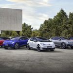 Fünf Elektroautos von Volkswagen stehen nebeneinander auf einem Parkplatz vor einem modernen Gebäude. Die Fahrzeuge sind in Rot, Blau, Weiß, Grau und Rot lackiert. Im Hintergrund sind Bäume und ein heller Himmel zu sehen.