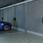 Tiefgarage eines Mehrfamilienhauses mit mehreren an der Betonwand montierten Wallboxen. Ein blaues Elektroauto steht an einem Ladeplatz und ist mit einer Ladestation verbunden. Entlang der Wand verläuft eine sichtbare Stromschiene mit mehreren Anschlüssen für Ladepunkte.