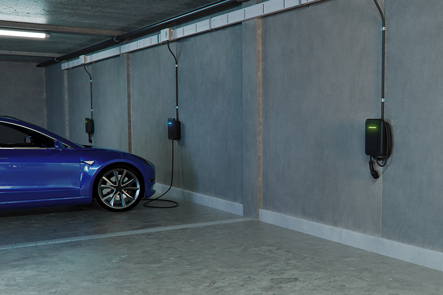 Tiefgarage eines Mehrfamilienhauses mit mehreren an der Betonwand montierten Wallboxen. Ein blaues Elektroauto steht an einem Ladeplatz und ist mit einer Ladestation verbunden. Entlang der Wand verläuft eine sichtbare Stromschiene mit mehreren Anschlüssen für Ladepunkte.
