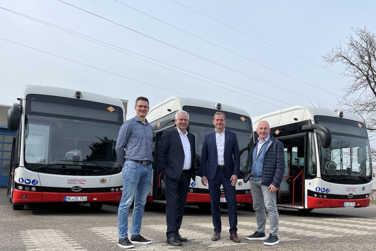 Vier Männer stehen vor drei neuen elektrischen Linienbussen der Stadtwerke Neuss auf einem Betriebshof und präsentieren die moderne, emissionsfreie Fahrzeugflotte.