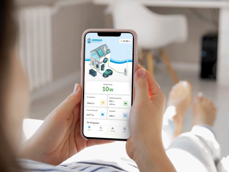 Eine Person hält ein Smartphone in der Hand und nutzt die App „Kilowatt.Käpt’n“ der WEMAG. Auf dem Display sind Energiedaten eines Haushalts zu sehen, darunter Stromproduktion, Verbrauch, Batteriespeicher und Einspeisung. Die App dient zur digitalen Steuerung von Eigenverbrauch und Energieflüssen im Zuhause.