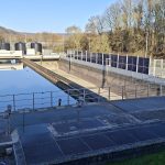 Blick über ein rechteckiges Becken der Kläranlage der Verbandsgemeindewerke Vordereifel mit ruhiger Wasseroberfläche. Daneben verläuft ein weiteres leeres Betonbecken mit technischen Schienen und Räumvorrichtungen. Im Vordergrund befinden sich Metallgitterstege und Geländer, rechts ein Zaun mit integrierten Solarmodulen. Im Hintergrund stehen mehrere schwarze Behälter sowie kahle Bäume unter blauem Himmel.