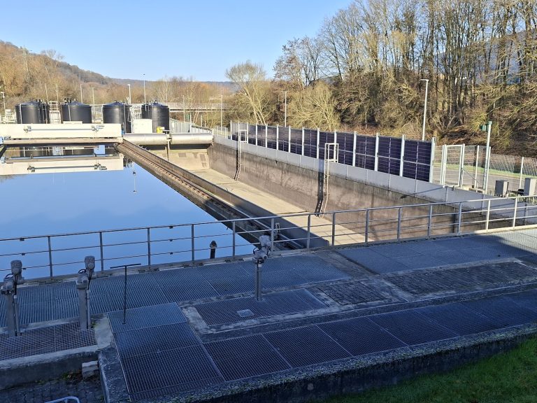 Blick über ein rechteckiges Becken der Kläranlage der Verbandsgemeindewerke Vordereifel mit ruhiger Wasseroberfläche. Daneben verläuft ein weiteres leeres Betonbecken mit technischen Schienen und Räumvorrichtungen. Im Vordergrund befinden sich Metallgitterstege und Geländer, rechts ein Zaun mit integrierten Solarmodulen. Im Hintergrund stehen mehrere schwarze Behälter sowie kahle Bäume unter blauem Himmel.