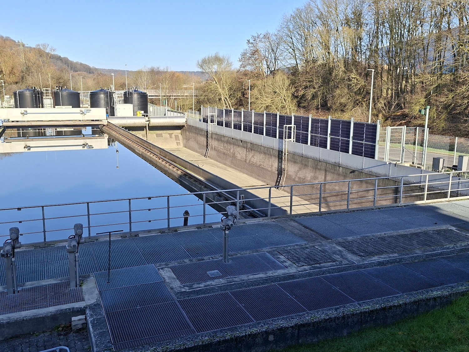Blick über ein rechteckiges Becken der Kläranlage der Verbandsgemeindewerke Vordereifel mit ruhiger Wasseroberfläche. Daneben verläuft ein weiteres leeres Betonbecken mit technischen Schienen und Räumvorrichtungen. Im Vordergrund befinden sich Metallgitterstege und Geländer, rechts ein Zaun mit integrierten Solarmodulen. Im Hintergrund stehen mehrere schwarze Behälter sowie kahle Bäume unter blauem Himmel.