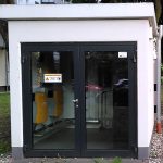 Vorderansicht eines kleinen, weißen Technikgebäudes des Quartierspeichers mit flachem Dach und verglaster Doppeltür. Im Inneren sind mehrere gelbe technische Geräte und Kabelinstallationen sichtbar. Das Gebäude steht auf einer Grünfläche zwischen Wohnhäusern und Bäumen.