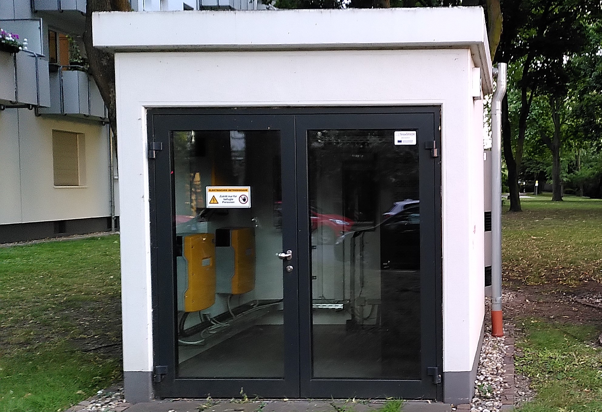 Vorderansicht eines kleinen, weißen Technikgebäudes des Quartierspeichers mit flachem Dach und verglaster Doppeltür. Im Inneren sind mehrere gelbe technische Geräte und Kabelinstallationen sichtbar. Das Gebäude steht auf einer Grünfläche zwischen Wohnhäusern und Bäumen.