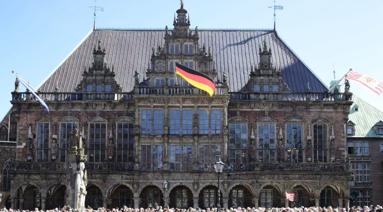 Frontansicht des Bremer Rathauses mit reich verzierter Backsteinfassade, großen Fenstern und historischem Giebel. Über dem Gebäude wehen die deutsche Flagge sowie weitere Fahnen. V