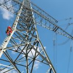 Arbeiter in Schutzkleidung und Helmen steigen an einem hohen Strommast aus Metall hinauf, während sie an Hochspannungsleitungen arbeiten; der Himmel ist blau mit wenigen Wolken.