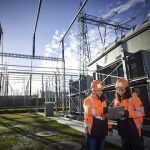 Zwei Mitarbeitende mit Schutzhelmen und Tablet in der Hand vor einem Umspannwerk mit Hochspannungsanlagen und Stromleitungen unter blauem Himmel.