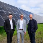 „Drei Männer stehen vor einer großen Freiflächen-Solaranlage auf einer grünen Wiese. Im Hintergrund erstrecken sich mehrere Reihen moderner Solarpaneele unter bewölktem Himmel. Zwei der Männer tragen Business-Outfits, der dritte eine dunkle Freizeitjacke. Die Aufnahme vermittelt den Eindruck eines offiziellen Termins oder einer Inbetriebnahme im Bereich erneuerbare Energien.“