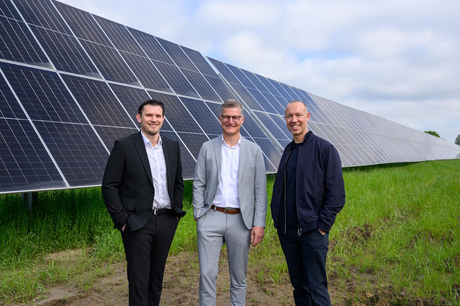 „Drei Männer stehen vor einer großen Freiflächen-Solaranlage auf einer grünen Wiese. Im Hintergrund erstrecken sich mehrere Reihen moderner Solarpaneele unter bewölktem Himmel. Zwei der Männer tragen Business-Outfits, der dritte eine dunkle Freizeitjacke. Die Aufnahme vermittelt den Eindruck eines offiziellen Termins oder einer Inbetriebnahme im Bereich erneuerbare Energien.“